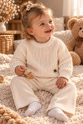 Baby trägt cremefarbenes Strickset mit Pullover und Hose