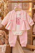 Rosafarbene Baby-Jacke mit weißem Blumenmuster und kleinen Schleifen, dazu heller Body mit Blumenmotiv und Hose