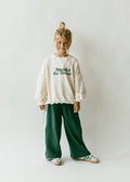 Mädchen im Kinder-Set mit cremefarbenem Sweatshirt mit „Snacks for Dinner“-Print und grüner weiten Hose, Studioshotaufnahme