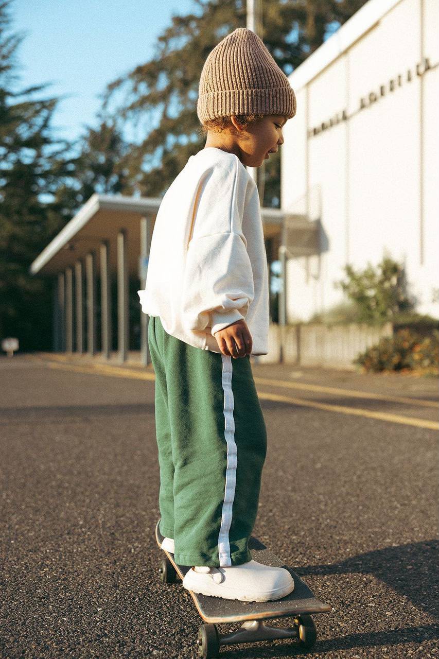 Kind von hinten auf dem Skateboard im cremefarbenen Sweatshirt und grüner weiten Hose mit weißem Seitenstreifen