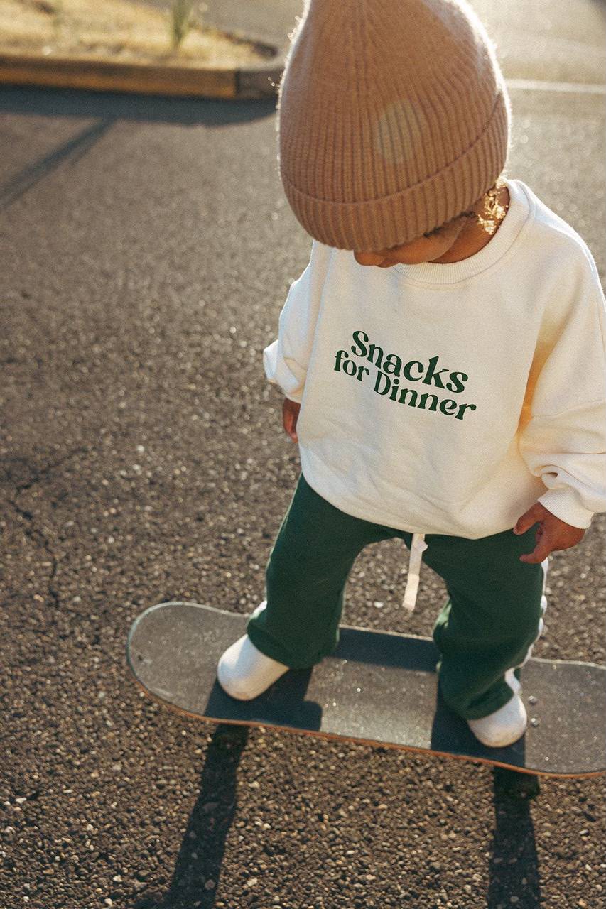 Kind im cremefarbenen Sweatshirt mit grünem „Snacks for Dinner“-Print und brauner Strickmütze
