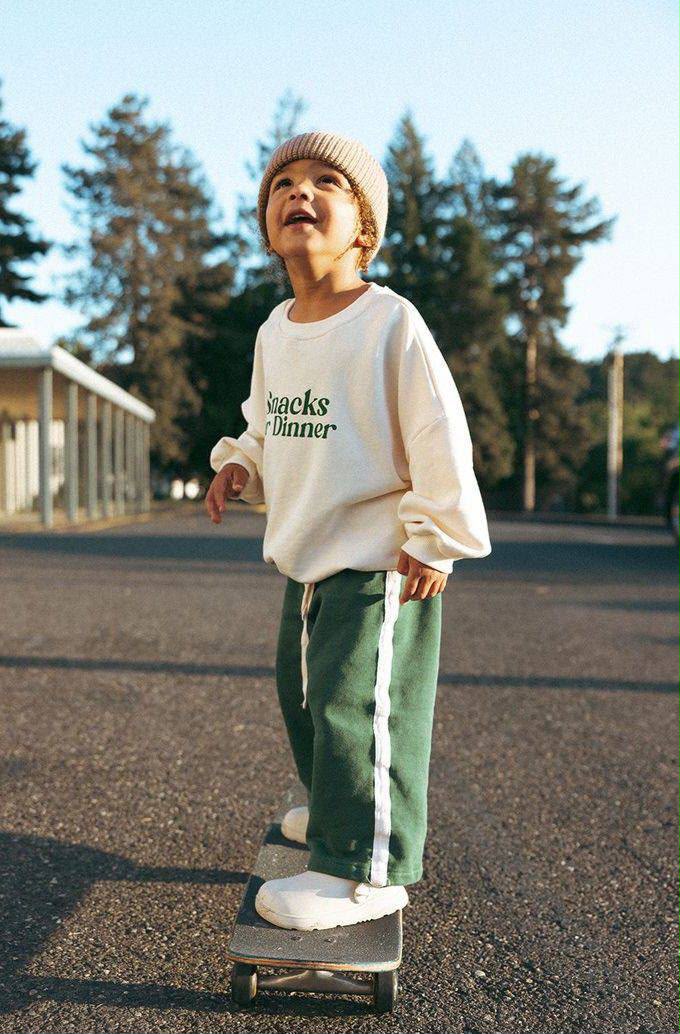 Kind auf dem Skateboard im Kinder-Set mit cremefarbenem Sweatshirt mit „Snacks for Dinner“-Print und grüner weiten Hose mit Kontraststreifen