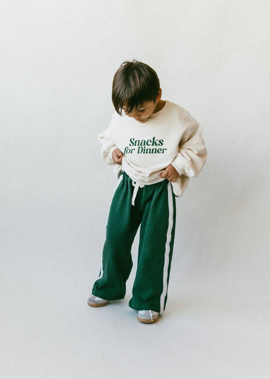 Junge im Kinder-Set mit cremefarbenem Sweatshirt mit „Snacks for Dinner“-Print und grüner weiten Hose, Studioshotaufnahme