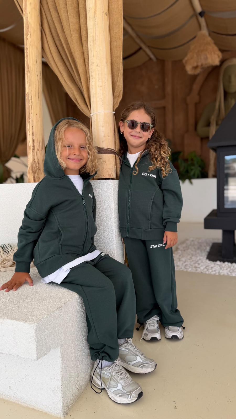 Zwei Kinder im dunkelgrünen Stay-Kind-Kinder-Set aus Zip-Hoodie-Jacke und weiter Hose auf einer Mauer sitzend