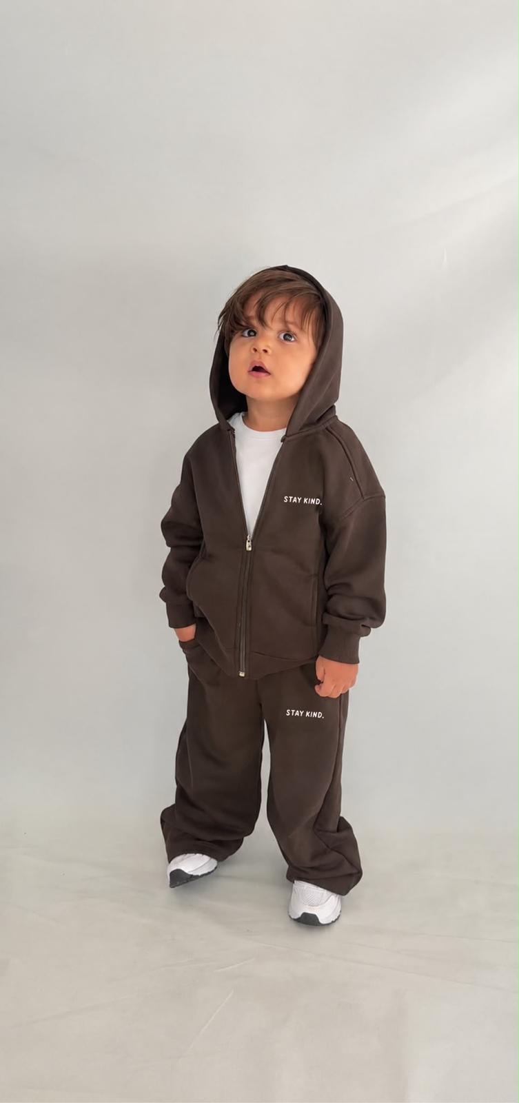  Kleinkind im dunkelbraunen Stay-Kind-Kinder-Set aus Zip-Hoodie-Jacke mit Kapuze und weiter Hose mit „Stay Kind“-Print