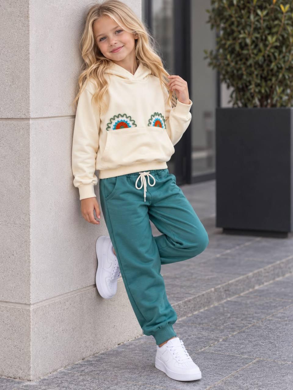 Mädchen lehnt an einer Wand im Rainbow Love Set mit cremefarbenem Regenbogen-Hoodie und petrolgrüner Hose.