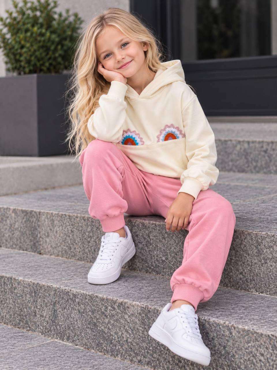 Mädchen sitzt auf einer Treppe im Rainbow Love Set mit cremefarbenem Hoodie, bunten Regenbogen-Applikationen und rosa Hose.