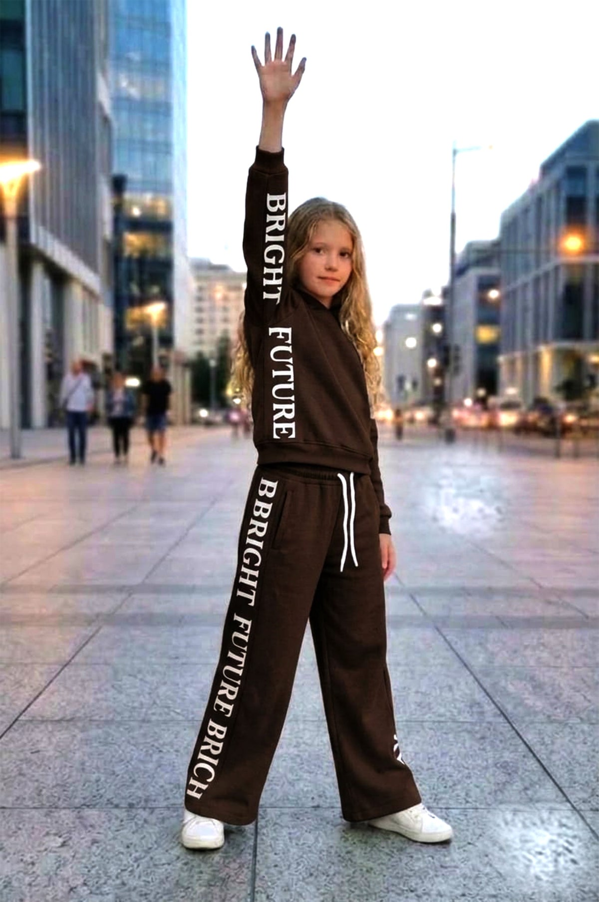 Bright Future Kids Set in Dunkelbraun: gefütterter Sweater und weite Hose mit Bright-Future-Schriftzug entlang des Ärmels und Hosenbeins, getragen mit weißen Sneakern.