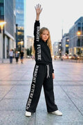 Bright Future Kids Set in Schwarz: gefütterter Sweater und weite Hose mit Bright-Future-Schriftzug entlang des Ärmels und Hosenbeins, getragen mit weißen Sneakern.