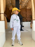 Stay Club Kids Set: Gesamtansicht des hellgrauen Sweat-Sets mit „Stay Club“-Print, getragen mit gelber Cap und Sneakern.