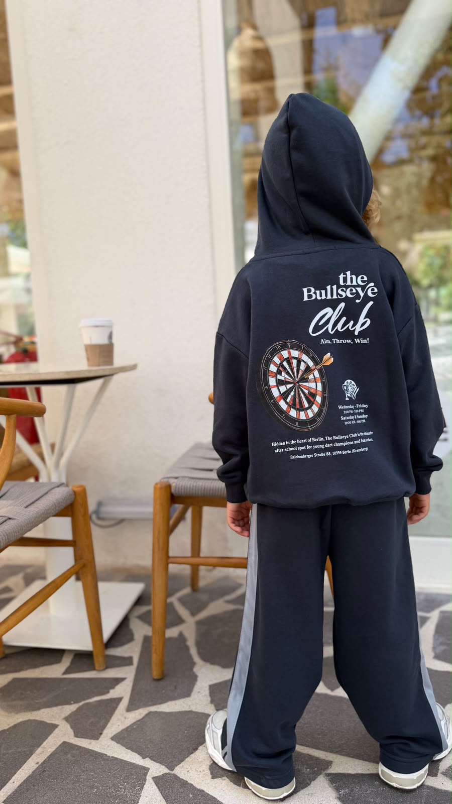 Kind zeigt den Rücken des Bullseye Club Hoodies mit Dartscheiben-Print und weiter Hose mit Streifen, Rückansicht