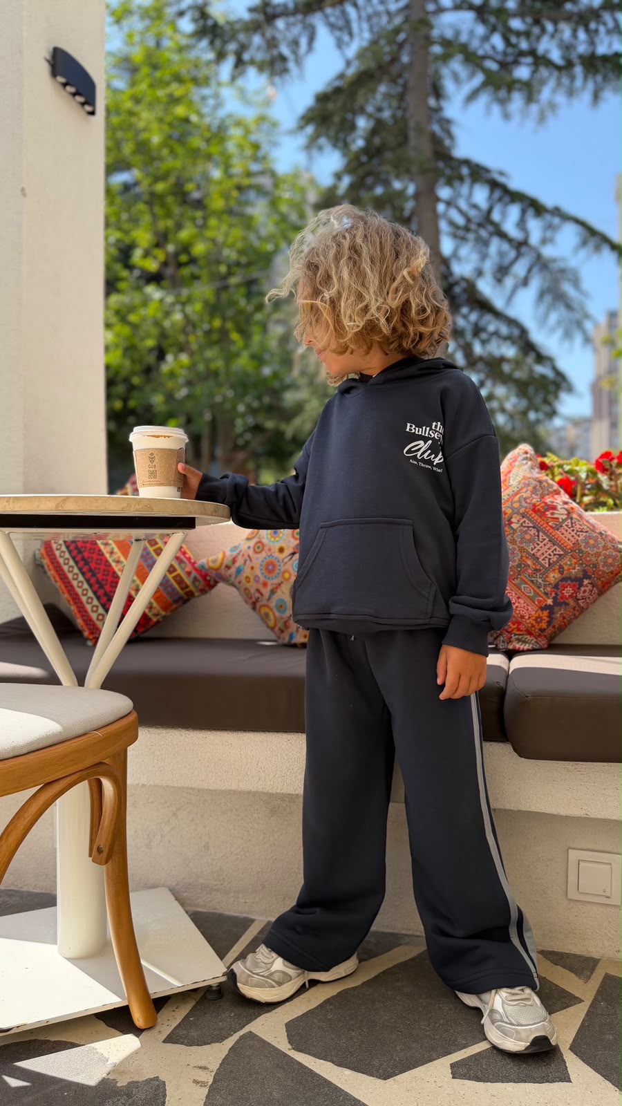 Kind im anthrazitfarbenen Bullseye Club Hoodie und weiter Hose mit Seitenstreifen am Cafétisch, Seitenansicht