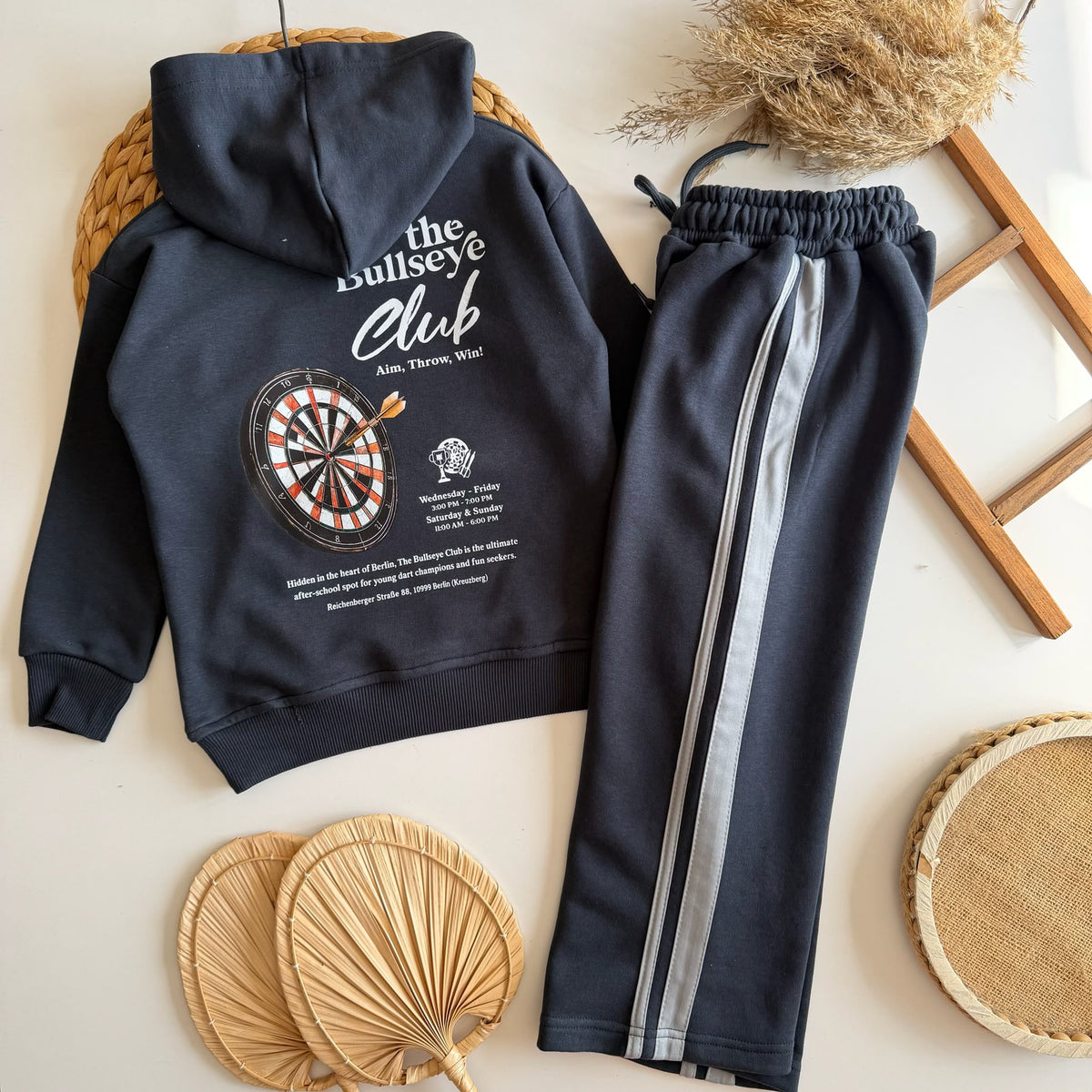 Bullseye Club Set flach liegend, anthrazitfarbener Hoodie mit Dartscheiben-Rückenprint und Hose mit hellen Seitenstreifen