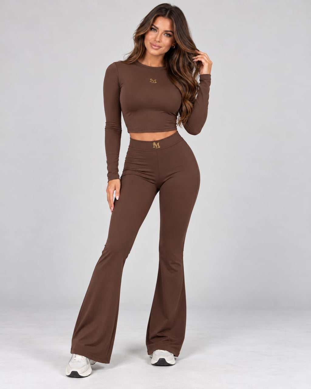 Model trägt ein braunes MILLAGRO Damen-Set mit figurbetontem Langarm-Crop-Top und High-Waist-Schlaghose zu weißen Sneakern.
