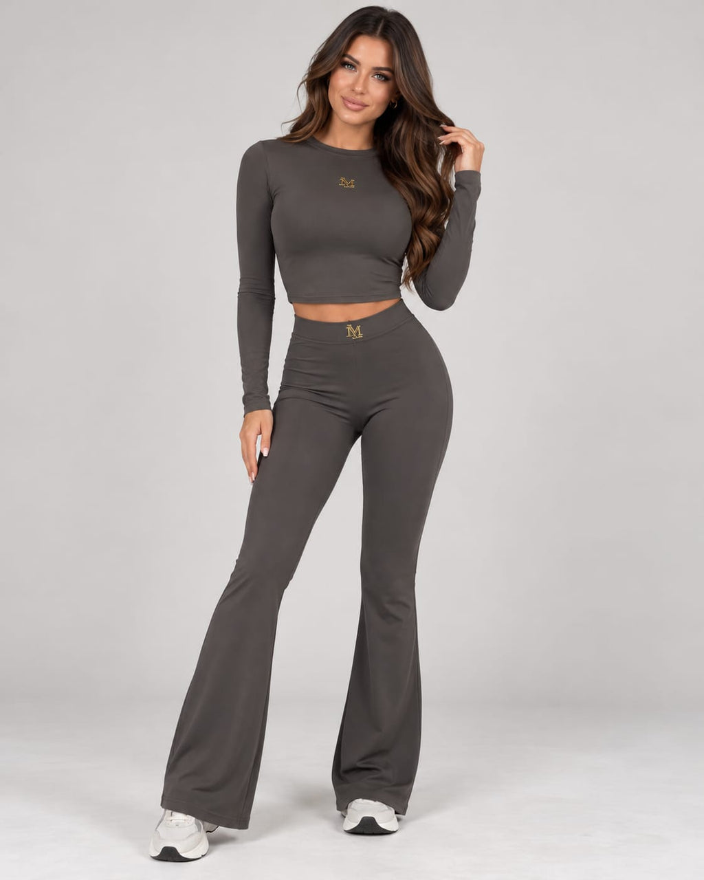 Model trägt ein graues MILLAGRO Damen-Set mit Langarm-Crop-Top, High-Waist-Schlaghose und goldfarbenen Logo-Details.