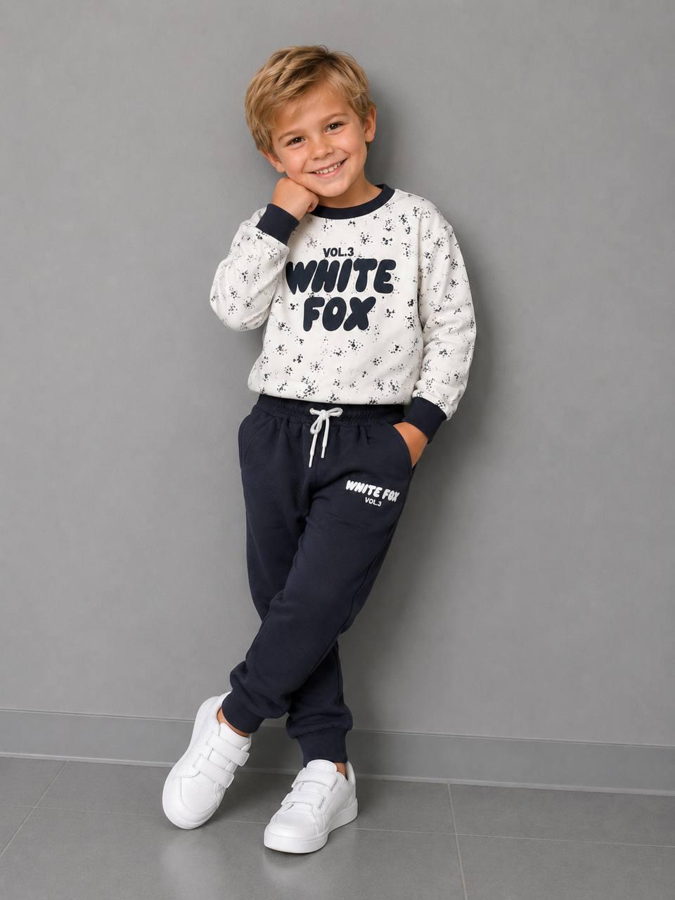Junge trägt ein Jungen-Set mit WHITE FOX Print, hellem gemustertem Sweatshirt mit schwarzen Bündchen und schwarzer Jogginghose.