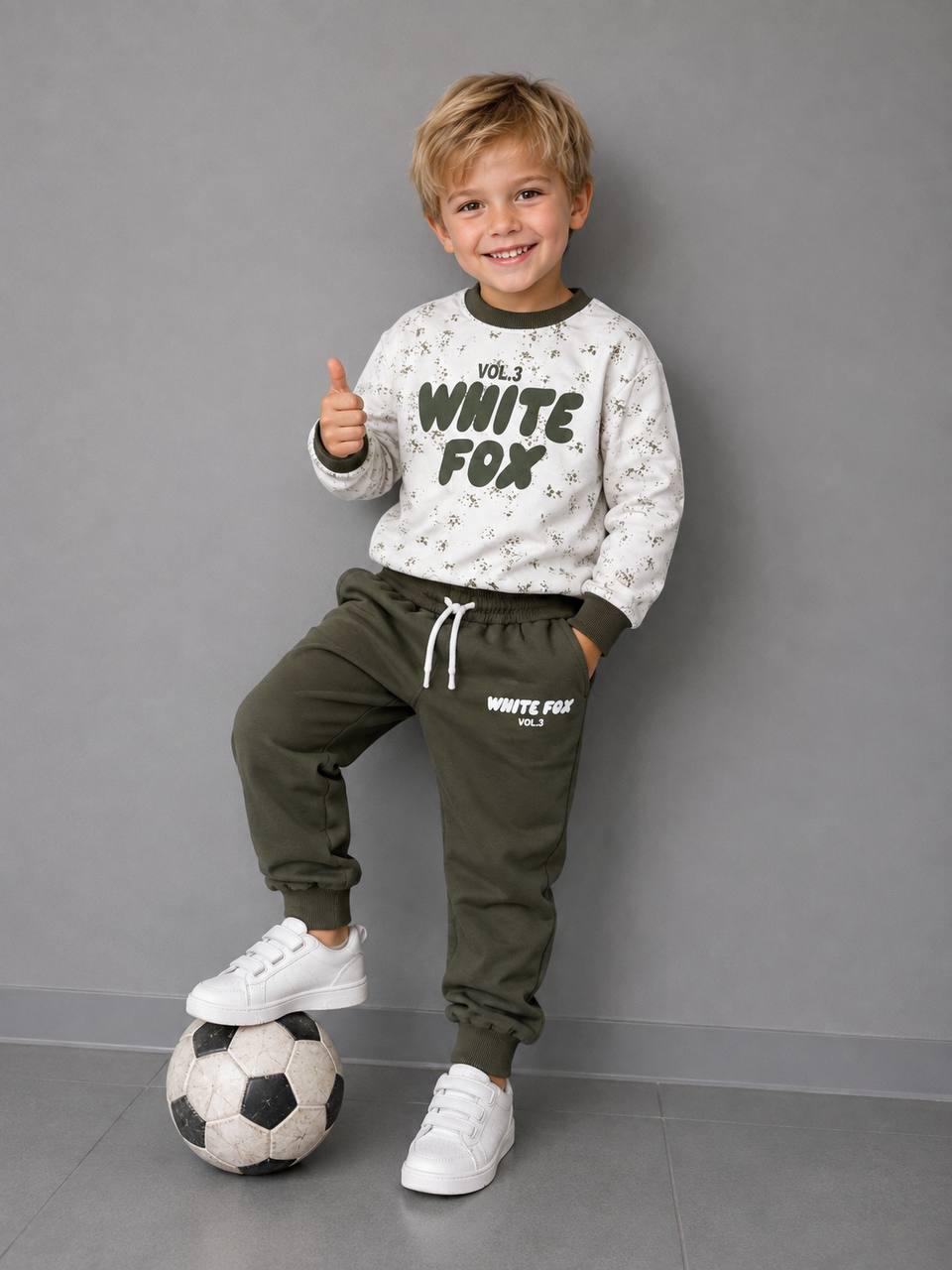 Junge trägt ein Jungen-Set mit WHITE FOX Print, hellem gemustertem Sweatshirt und khakifarbener Jogginghose, mit einem Fuß auf einem Fußball.