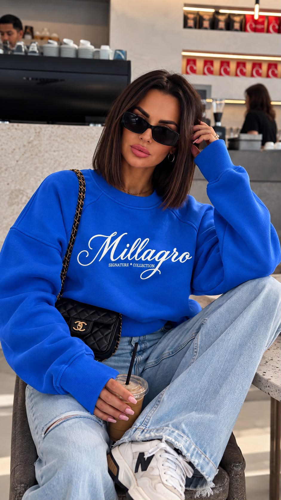 MILLAGRO Damen-Sweatshirt in Royalblau mit weißer Logo-Stickerei, an einem sitzenden Model mit Jeans und Sneakern.
