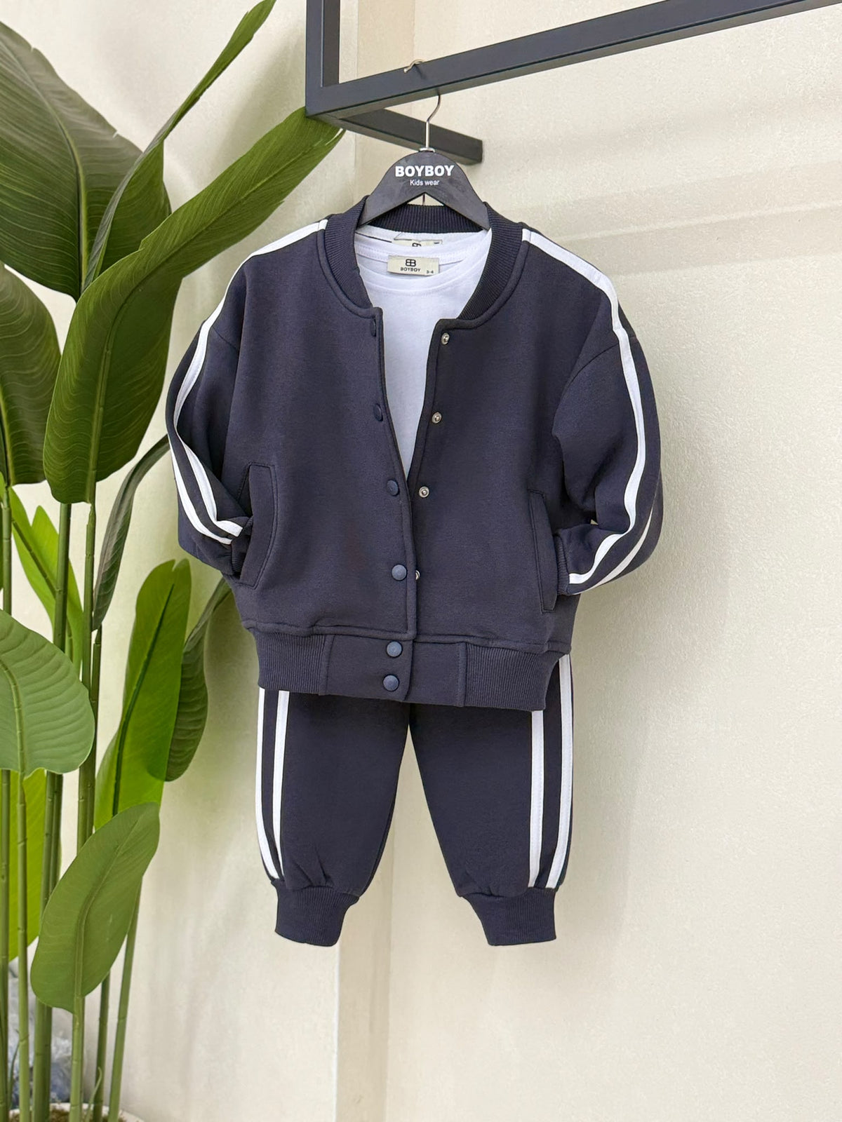 Dunkelgraues Kinder-Set mit Druckknopfjacke, weißem T-Shirt und Jogginghose mit weißen Seitenstreifen auf einem Kleiderbügel