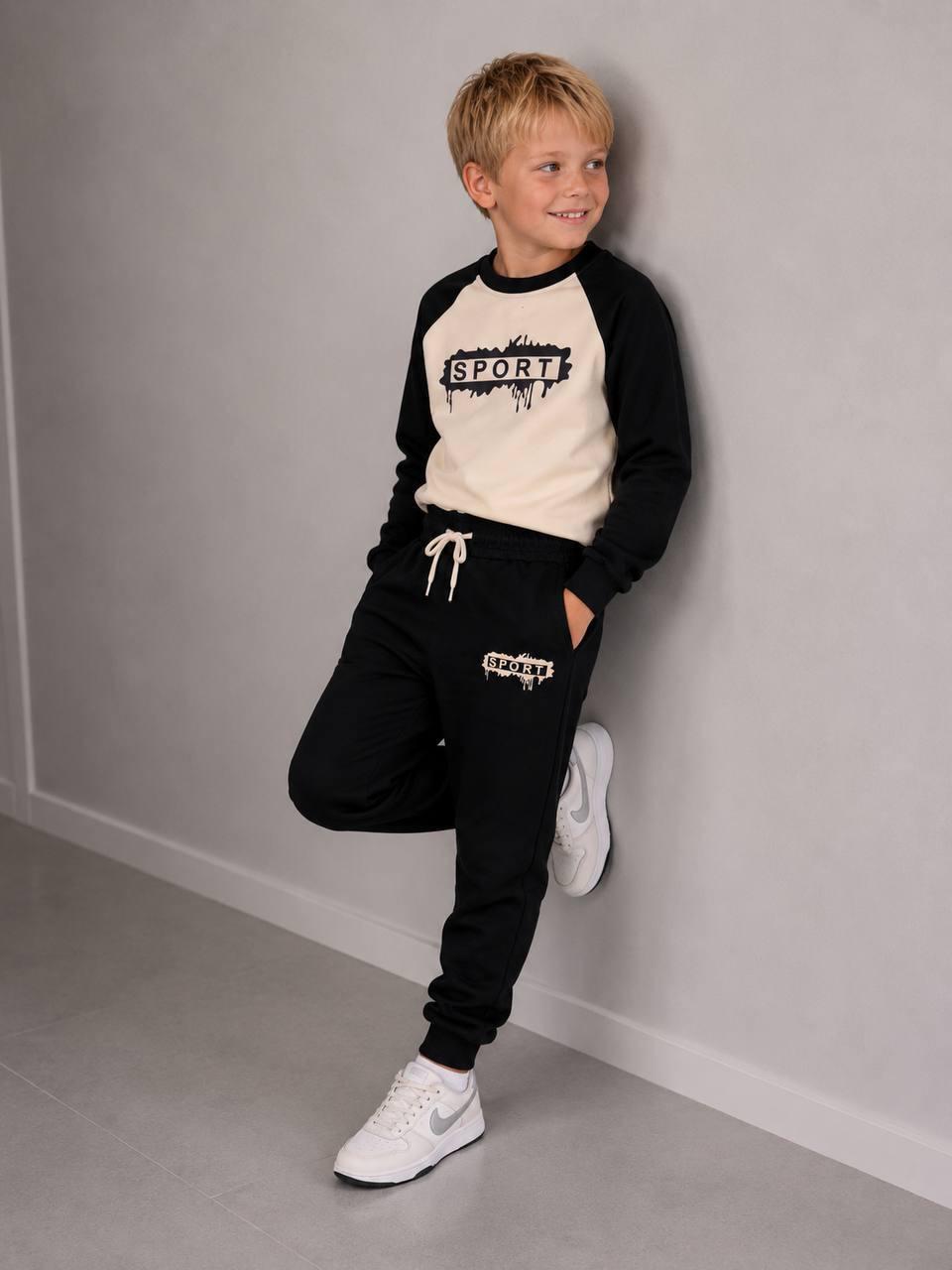 Junge trägt das schwarz-cremefarbene Happy Street Sweatset mit SPORT-Print und Jogginghose, an eine Wand gelehnt.