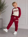 Junge zeigt das bordeauxrot-cremefarbene Happy Street Set mit SPORT-Print auf Sweatshirt und Jogginghose.