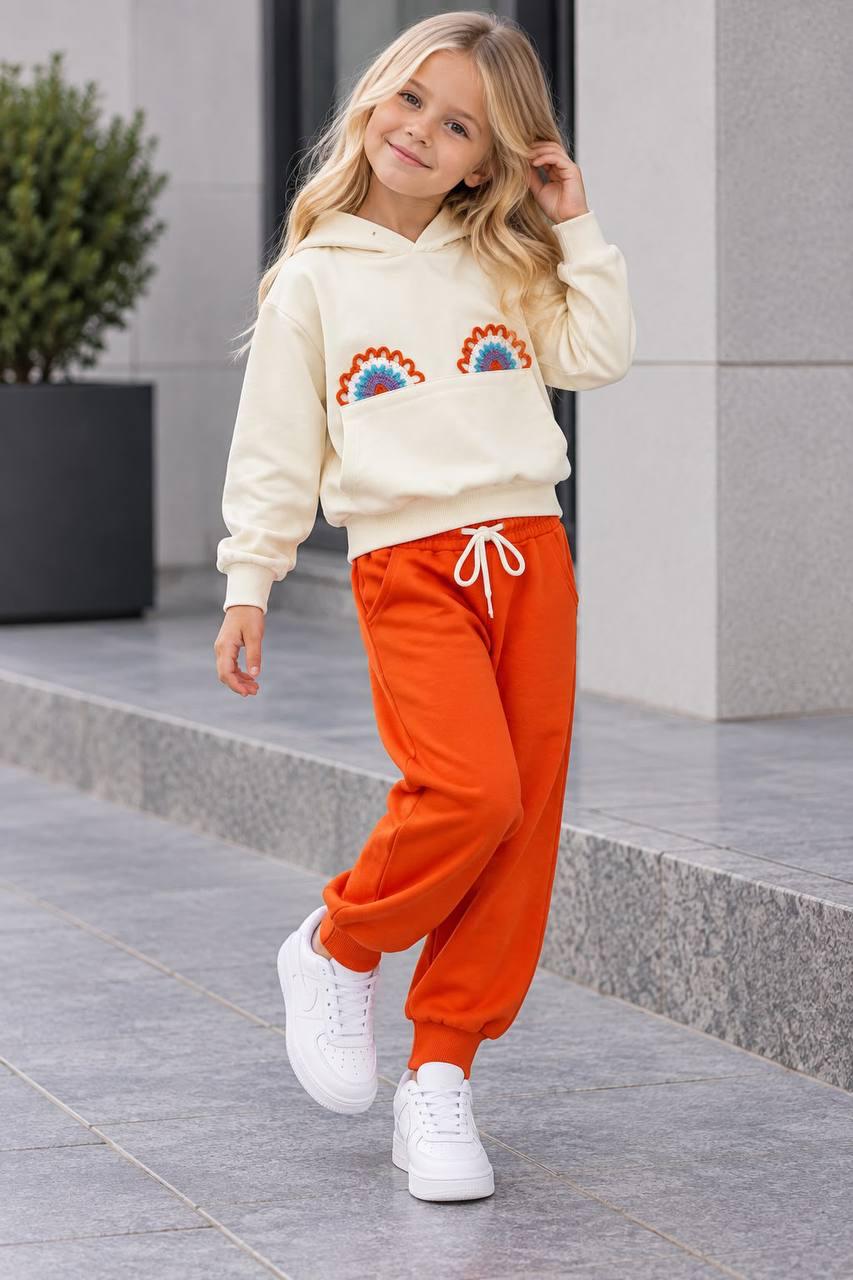 Mädchen zeigt das Rainbow Love Set mit cremefarbenem Hoodie, bunten Regenbogen-Applikationen und orangefarbener Hose mit Kordelzug.