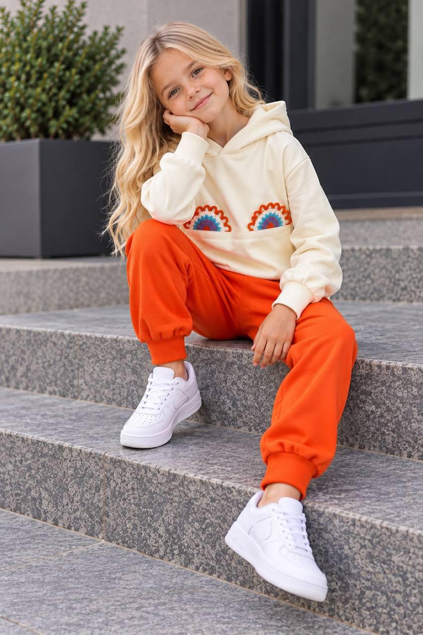 Mädchen sitzt auf einer Treppe im Rainbow Love Set mit cremefarbenem Regenbogen-Hoodie und orangefarbener Hose.