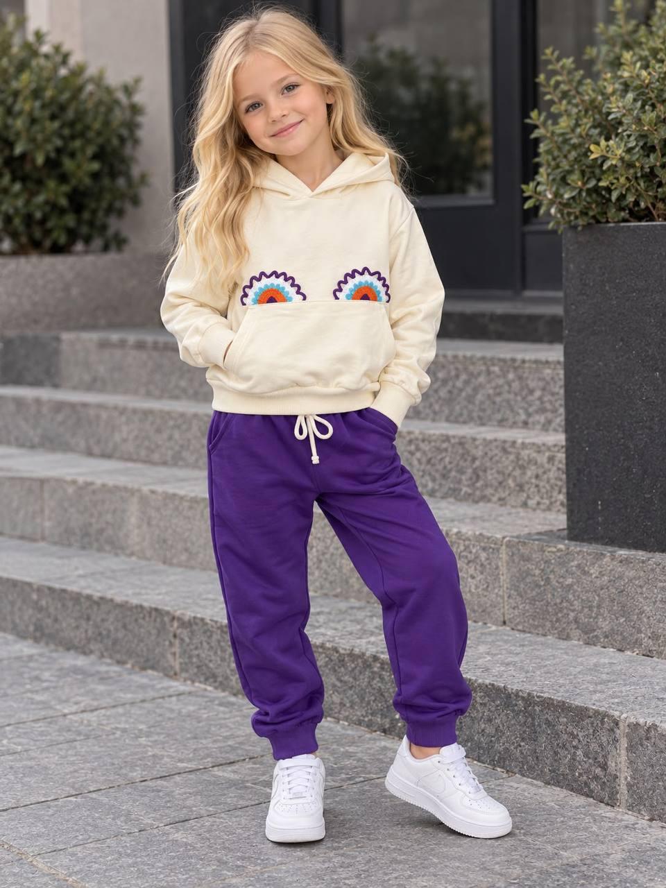 Mädchen trägt das Rainbow Love Set mit cremefarbenem Regenbogen-Hoodie und dunkellila Hose zu weißen Sneakern.