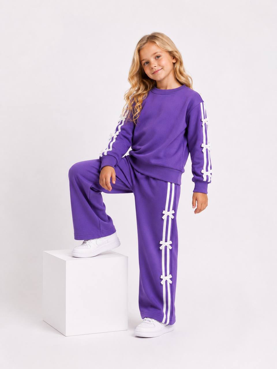 Mädchen trägt ein lila zweiteiliges Trainingsset mit Sweatshirt und weiter Hose sowie weißen Streifen und Schleifen-Details