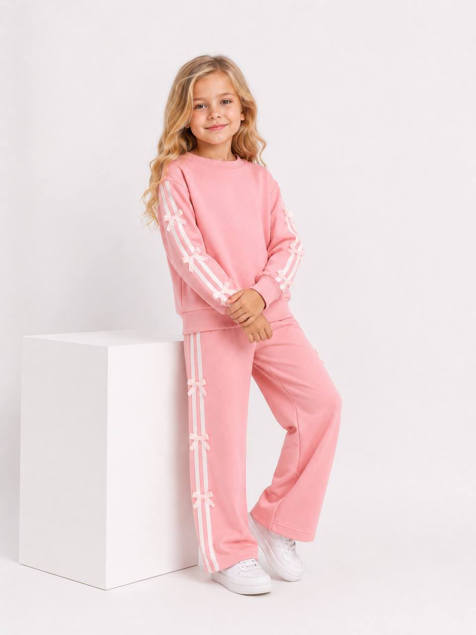Mädchen trägt ein rosafarbenes zweiteiliges Trainingsset mit Sweatshirt und weiter Hose sowie weißen Streifen und Schleifen-Details