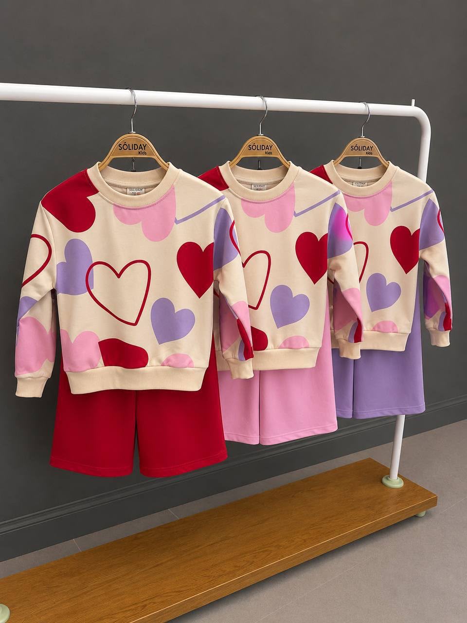 Zweiteiliges Mädchen-Set mit cremefarbenem Sweatshirt mit buntem Herz-Print und Weithosen in Rosa, Lila und Rot auf einer Kleiderstange