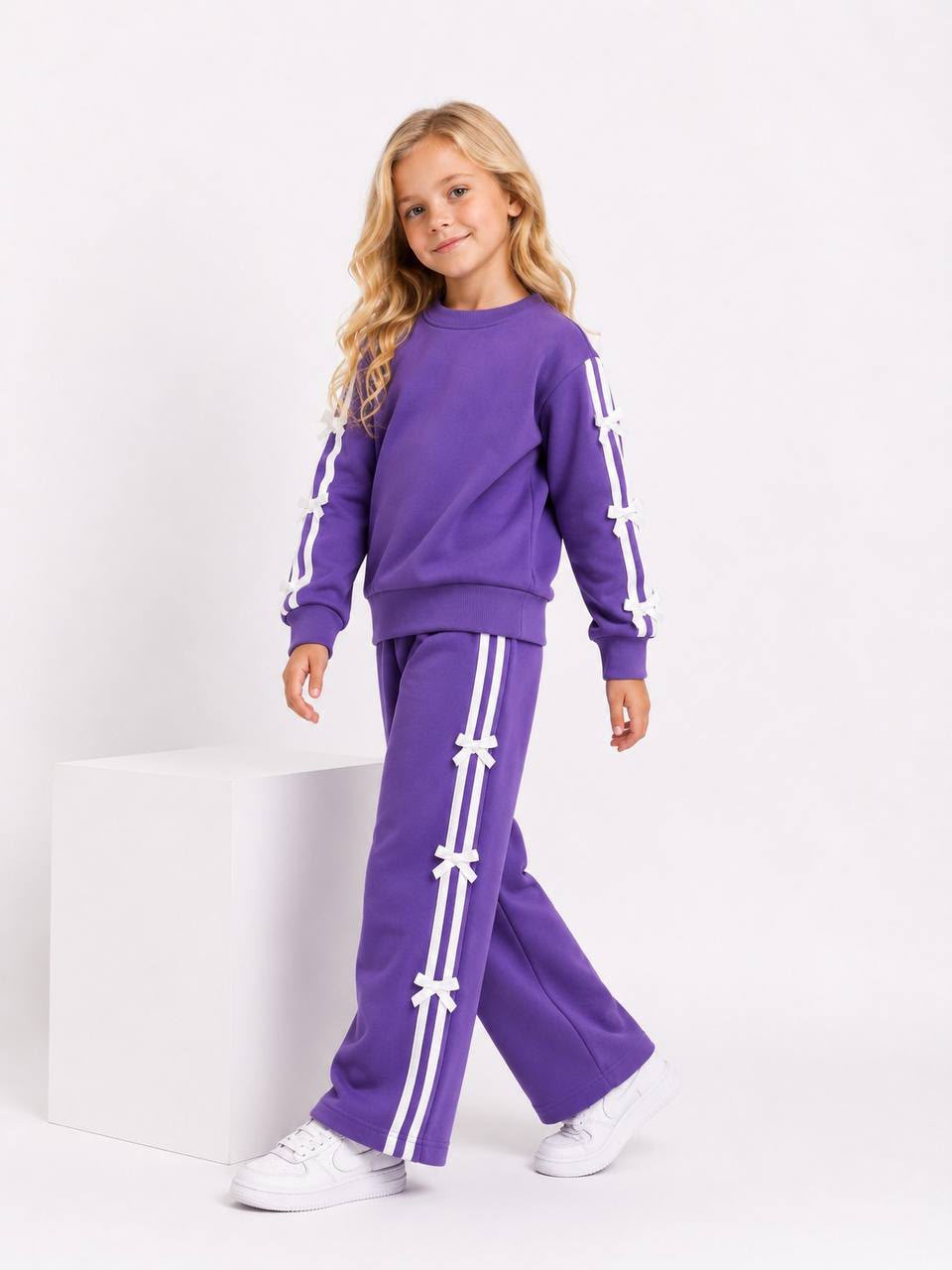 Mädchen trägt ein lila Trainingsset mit langärmligem Sweatshirt und weiter Hose mit weißen Streifen und Schleifen-Details